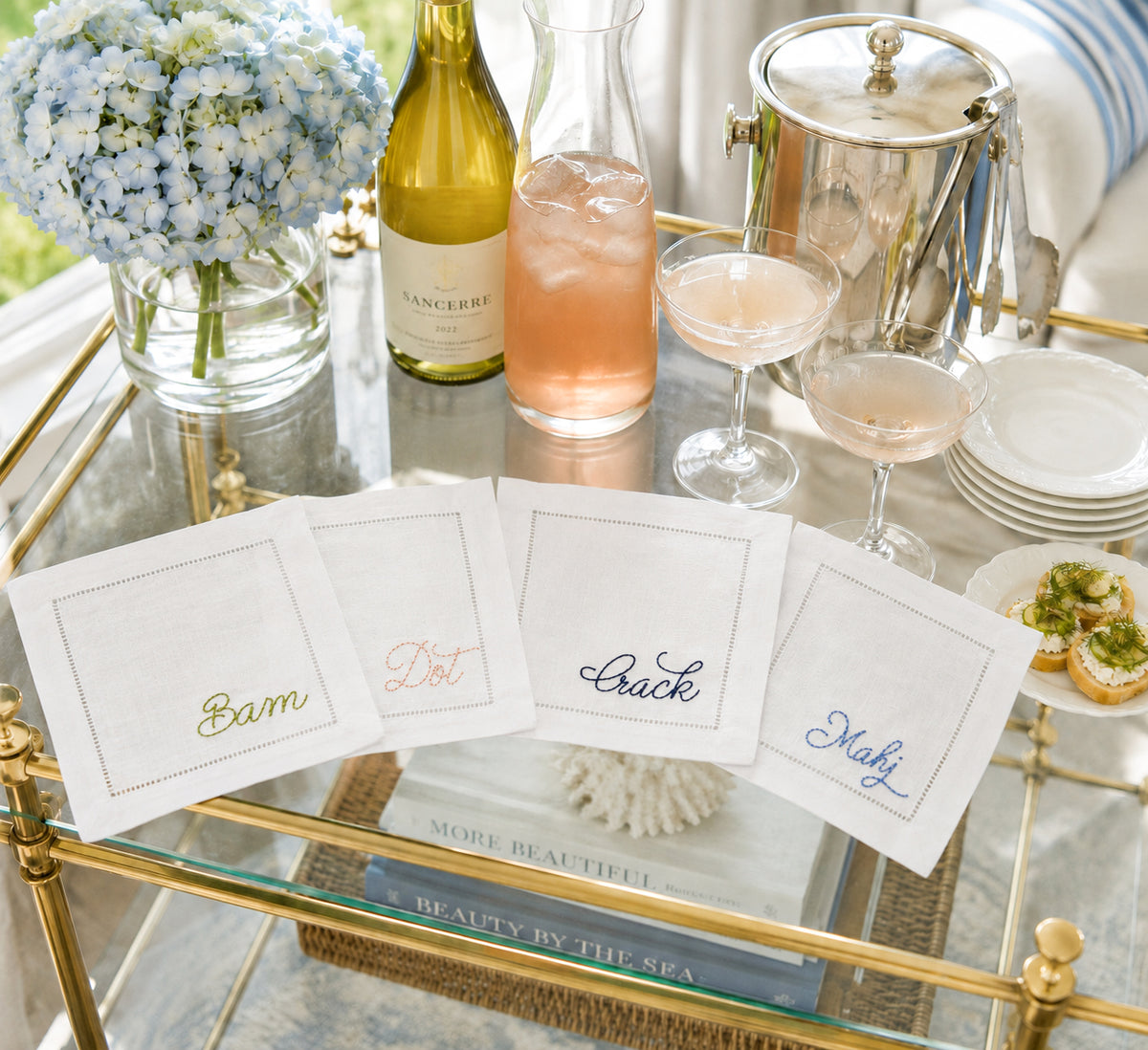 Four white embroidered linen cocktail napkins with the words (Bam, Dot, Crack, Mahj) are displayed on a glass-top serving cart alongside a wine bottle, glasses, flowers, and a metal ice bucket in a bright, styled setting.