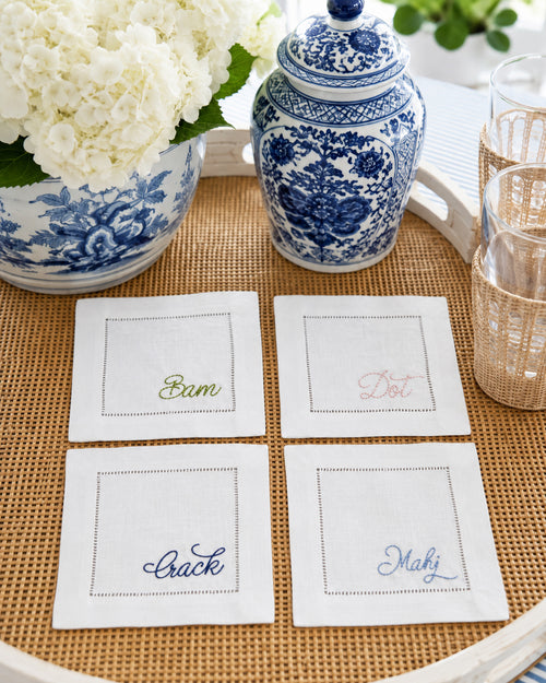 Four square white linen cocktail napkins with embroidered mahjong sayings including: bam, dot, crack, mahj, arranged on a woven tray alongside blue-and-white ceramic dishes and glassware.