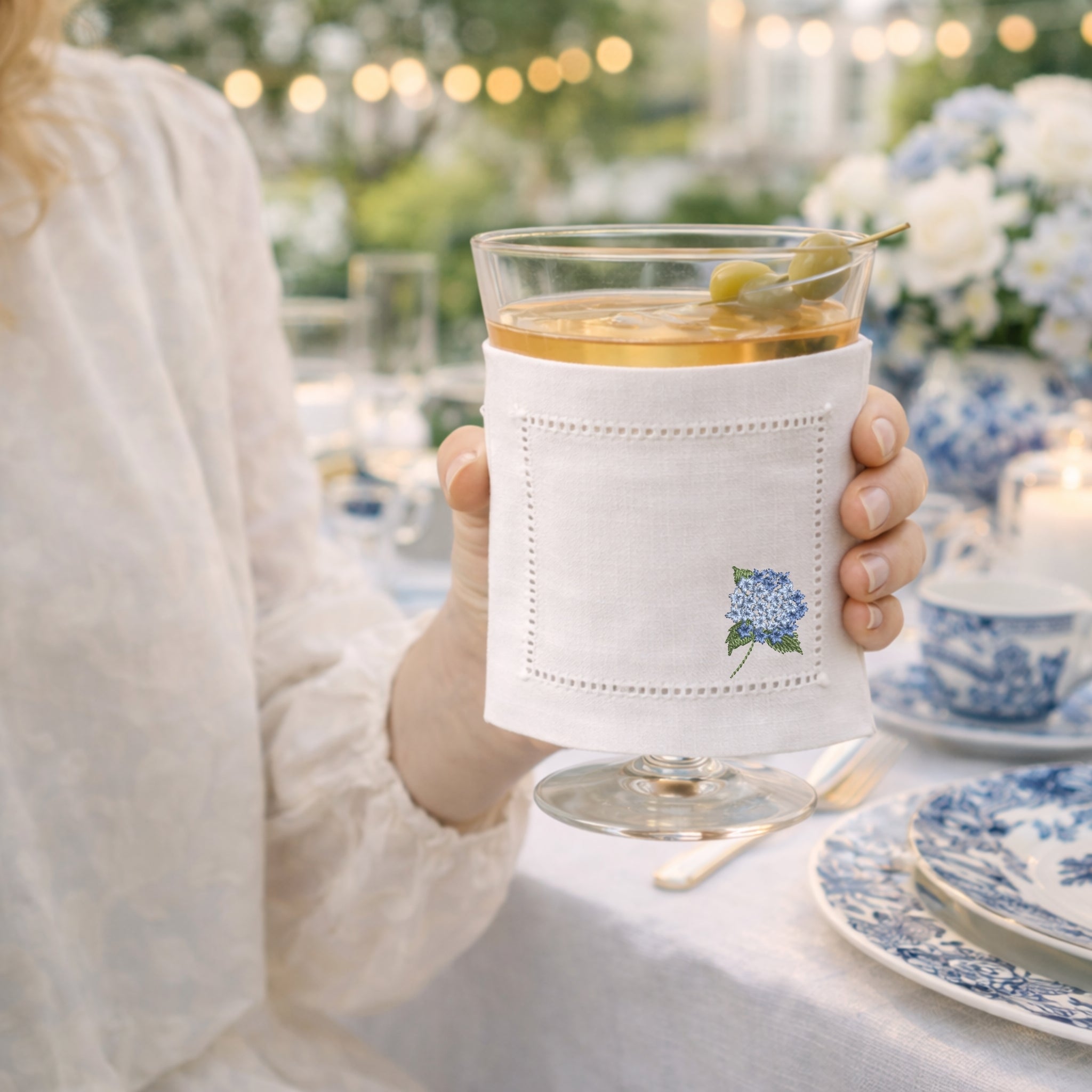 Person holding a glass with a white sleeve featuring a hydrangea floral design cocktail napkin, set against a blurred outdoor dining scene.