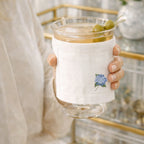 Hand holding a cocktail glass with a white sleeve featuring a blue hydrangea flower design, against a blurred indoor background.