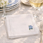 White embroidered napkin with a floral hydrangea design on the bottom right corner on a table setting with wine glasses and cutlery.