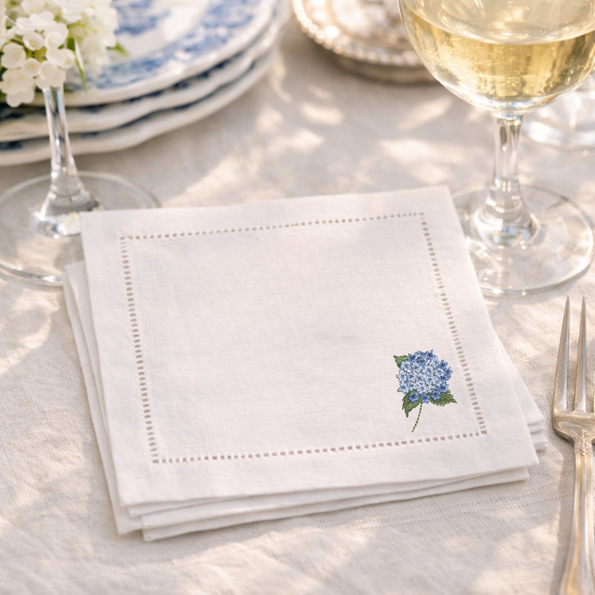 White embroidered napkin with a floral hydrangea design on the bottom right corner on a table setting with wine glasses and cutlery.