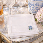 White napkin with embroidered hydrangea flower design on a decorative bar cart setting with glasses and flowers.