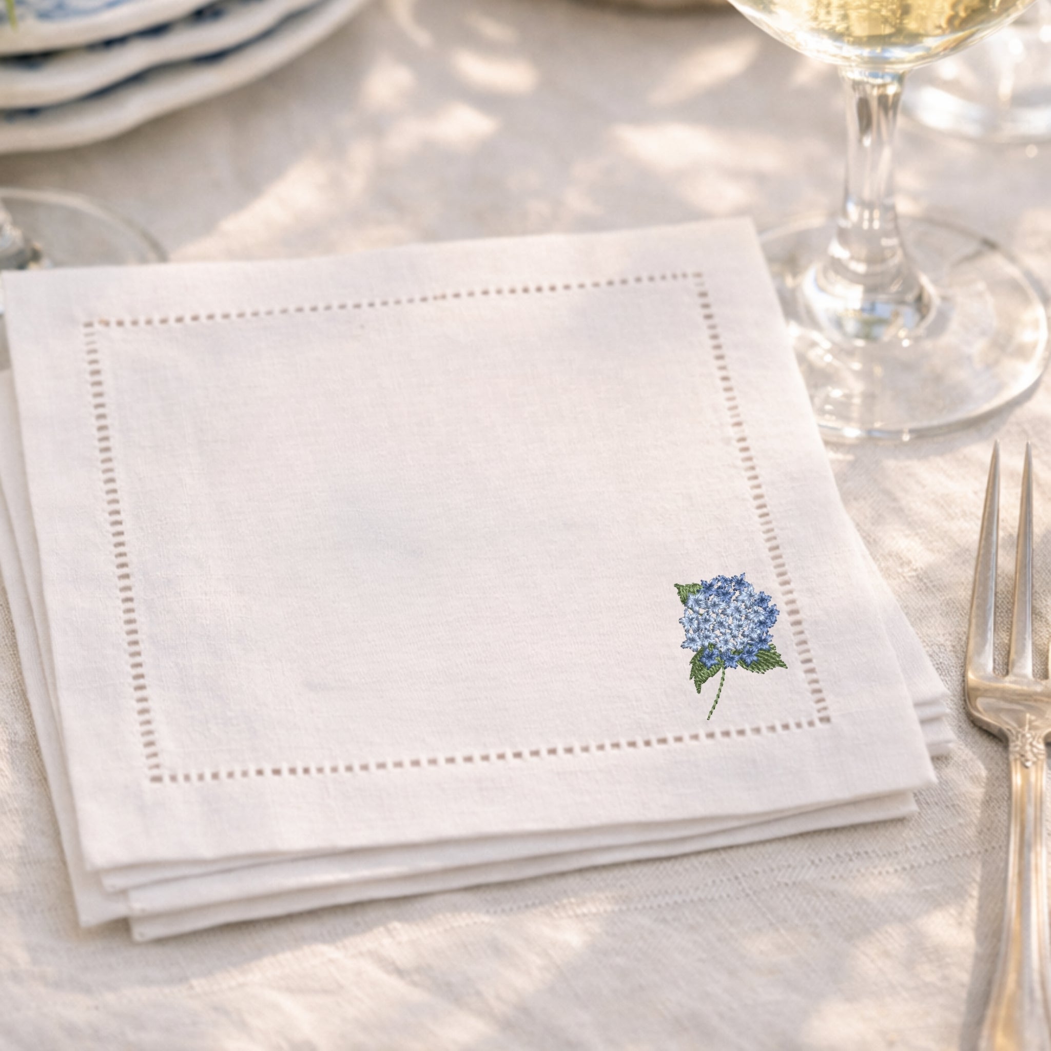 White napkin with embroidered blue flower design on a table setting with cutlery and glasses.