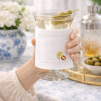 Hand holding a cocktail glass with olives, wrapped in a white napkin with embroidery, on a blurred background.