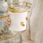 Glass with a white sleeve featuring a embroidered pomegranate design, held by a person in a blurred indoor setting.