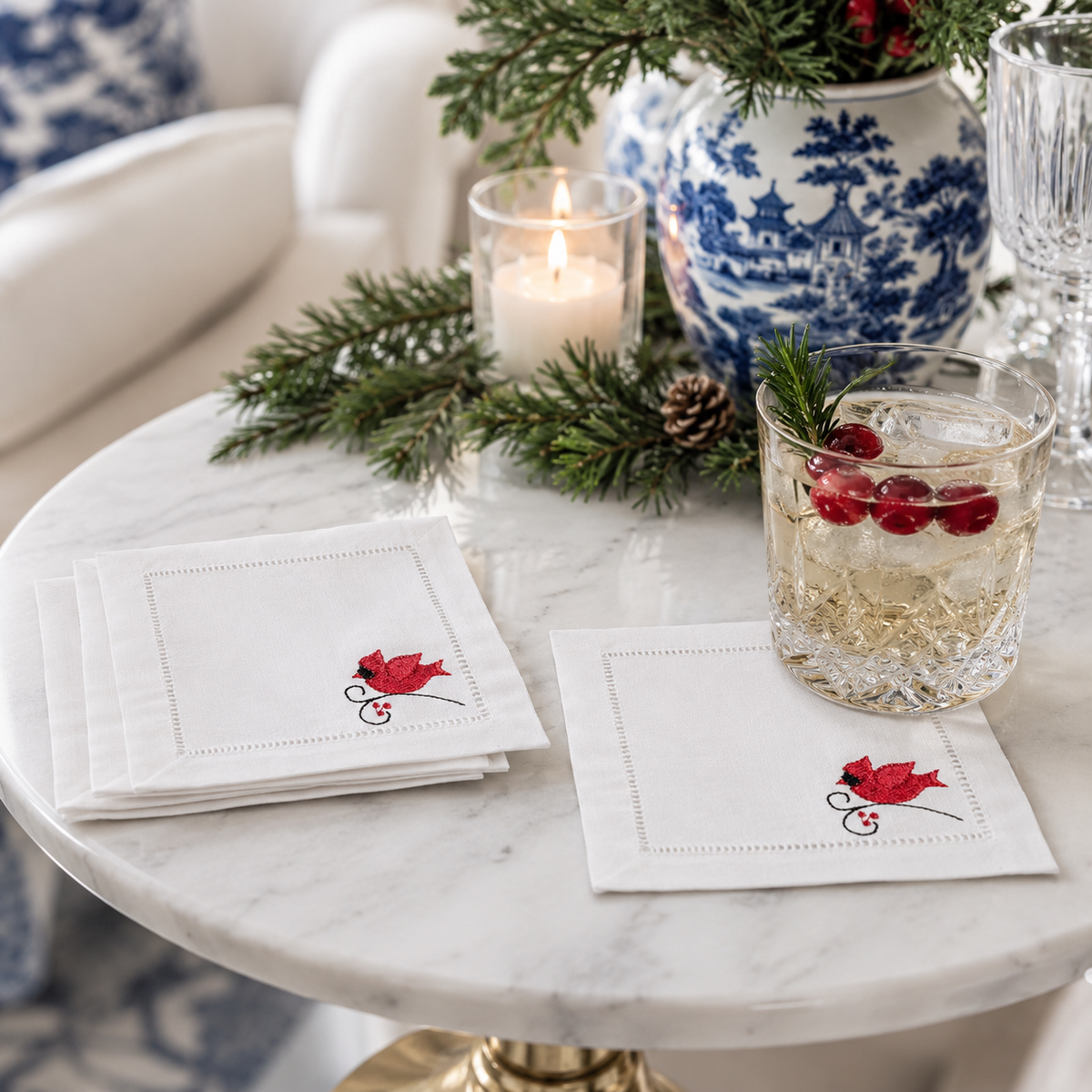 White cocktail napkins with a red cardinal in the bottom right, a hemstitch border and holiday decor.