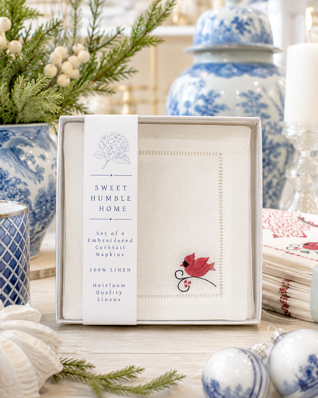 A boxed set of four embroidered cocktail napkins from Sweet Humble Home, displayed on a wooden table with holiday decor and blue-and-white ceramic vases in the background. The napkins are white linen with delicate hemstitched borders and a small red cardinal embroidered motif.