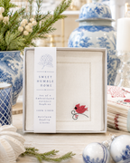 A boxed set of four embroidered cocktail napkins from Sweet Humble Home, displayed on a wooden table with holiday decor and blue-and-white ceramic vases in the background. The napkins are white linen with delicate hemstitched borders and a small red cardinal embroidered motif.