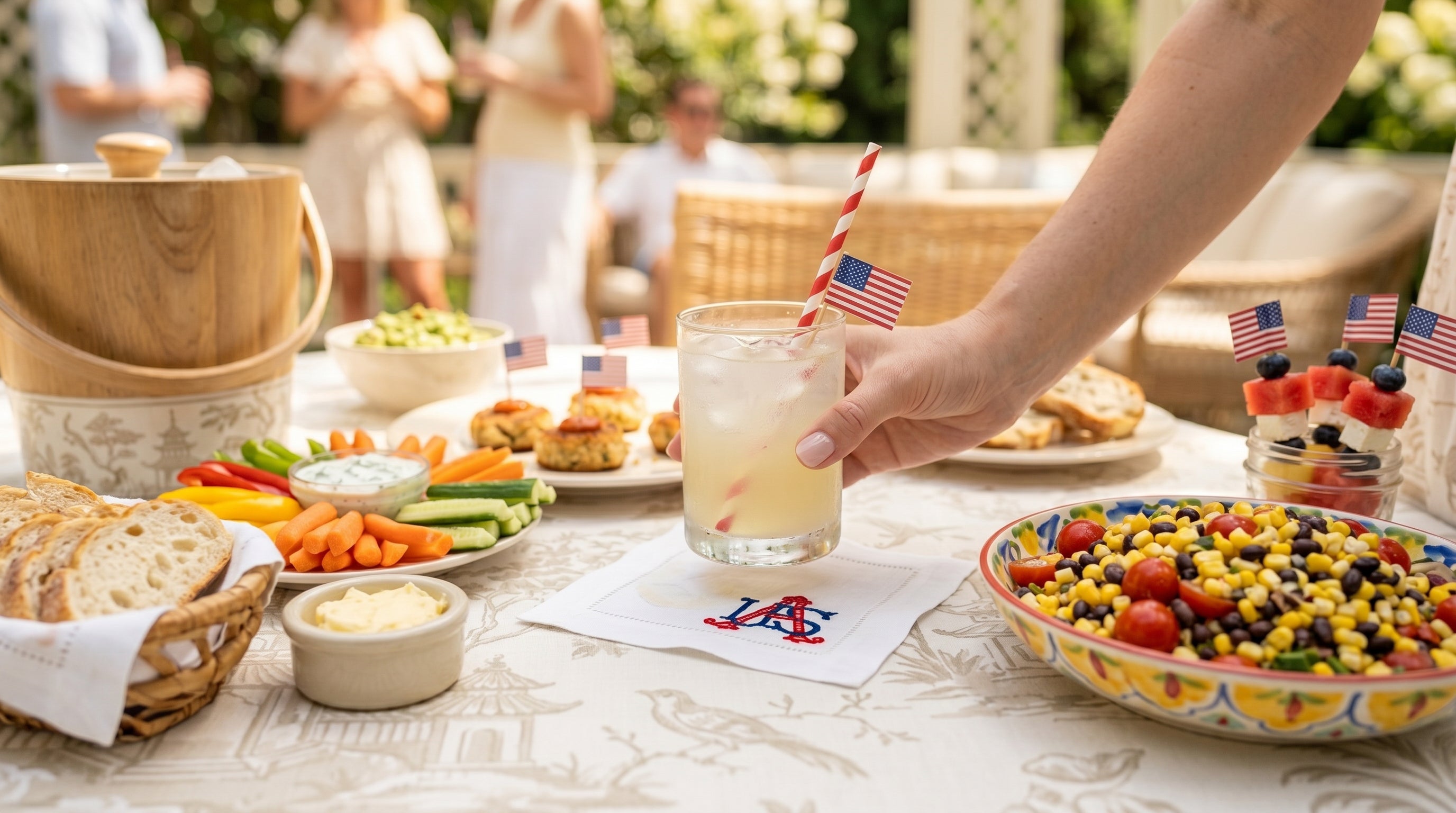 Outdoor setting with a table full of food and drinks, including a hand holding a drink with a straw. The person is putting a cocktail down onto a white linen table napkin embroidered with USA in the bottom corner in red and blue.