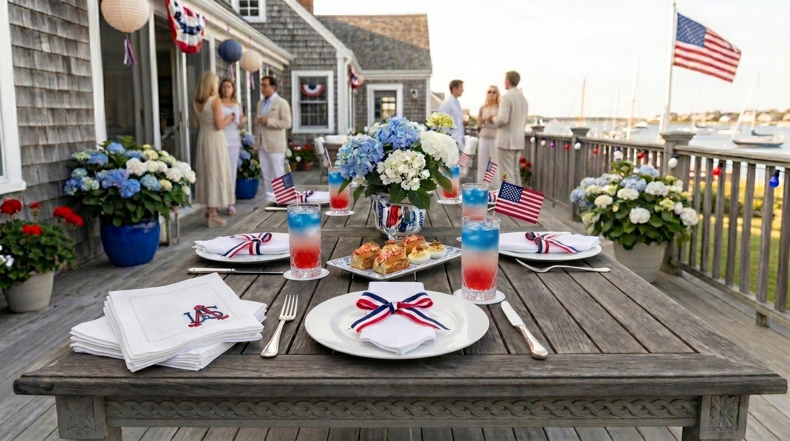 Outdoor table setting with American flag decorations on a deck. There are white place settings, red, white, and blue drinks, embroidered cocktail napkins embroidered with USA in the bottom right corner. The scene is overlooking water and boats.