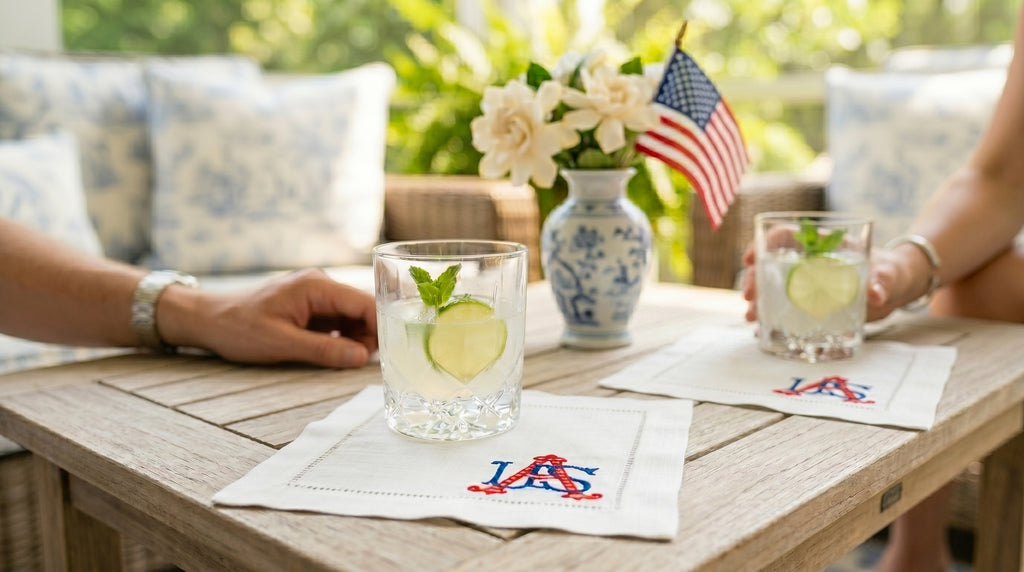 Two glasses with lime wedges on a table with embroidered cocktail napkins with USA embroidered in the bottom right corner, American flag, and flowers.