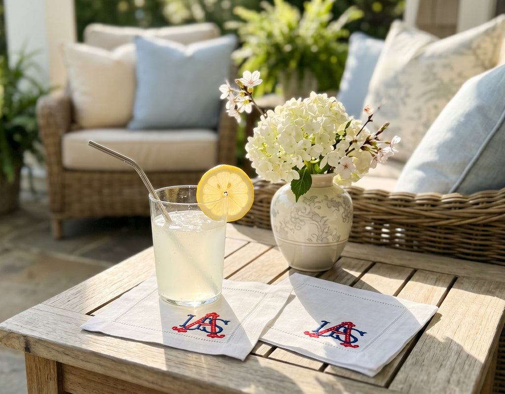 White linen cocktail napkins embroidered with USA in the bottom, right corner in blue and red sitting on a table with a vase and glass of lemonade outside.