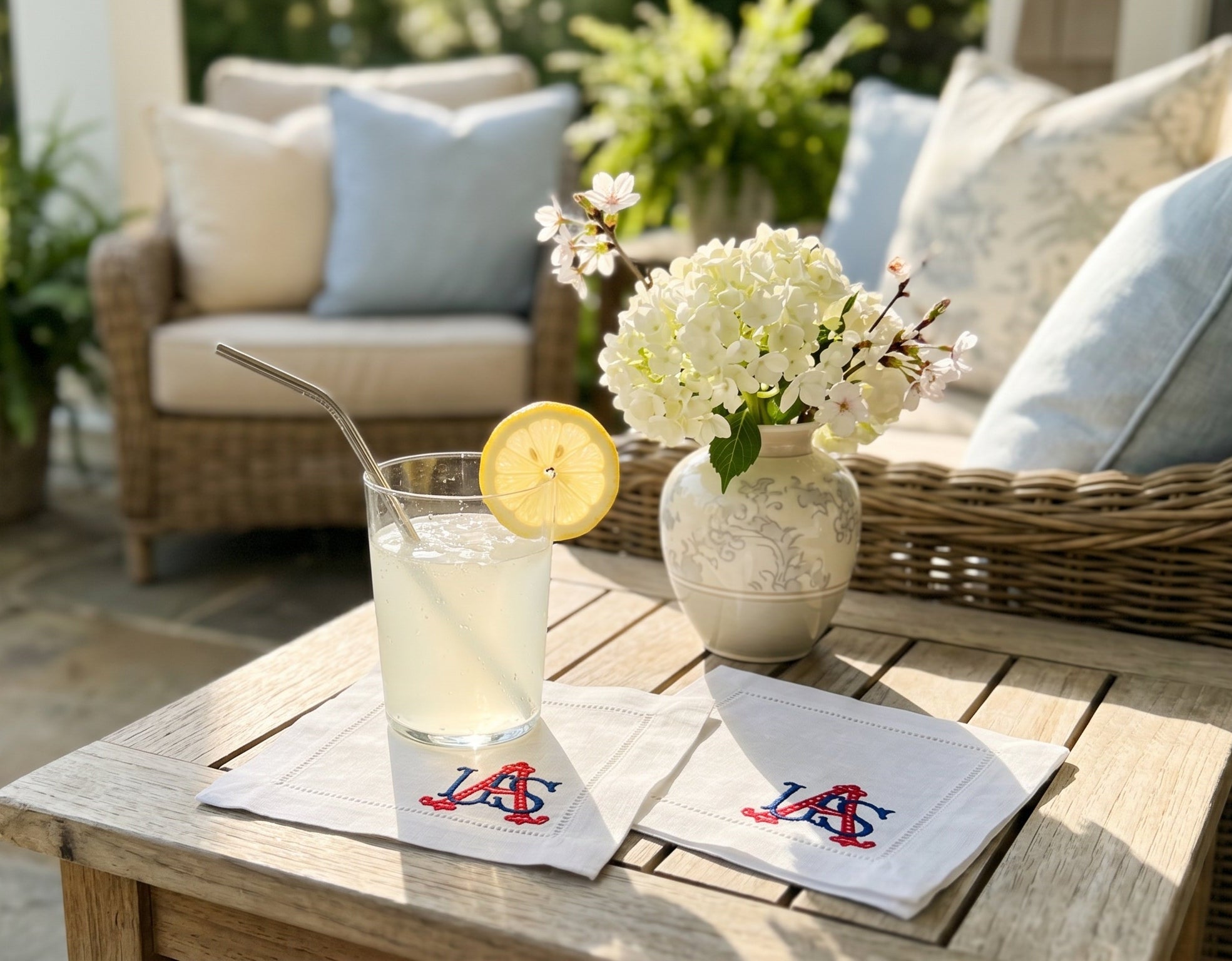 White linen cocktail napkins embroidered with USA in the bottom, right corner in blue and red sitting on a table with a vase and glass of lemonade outside.