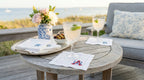 Outdoor setting with a wooden table, glasses of lemonade, and flowers on a deck. There are white linen embroidered cocktail napkins with hydrangeas and one with USA embroidered in the bottom right corner.