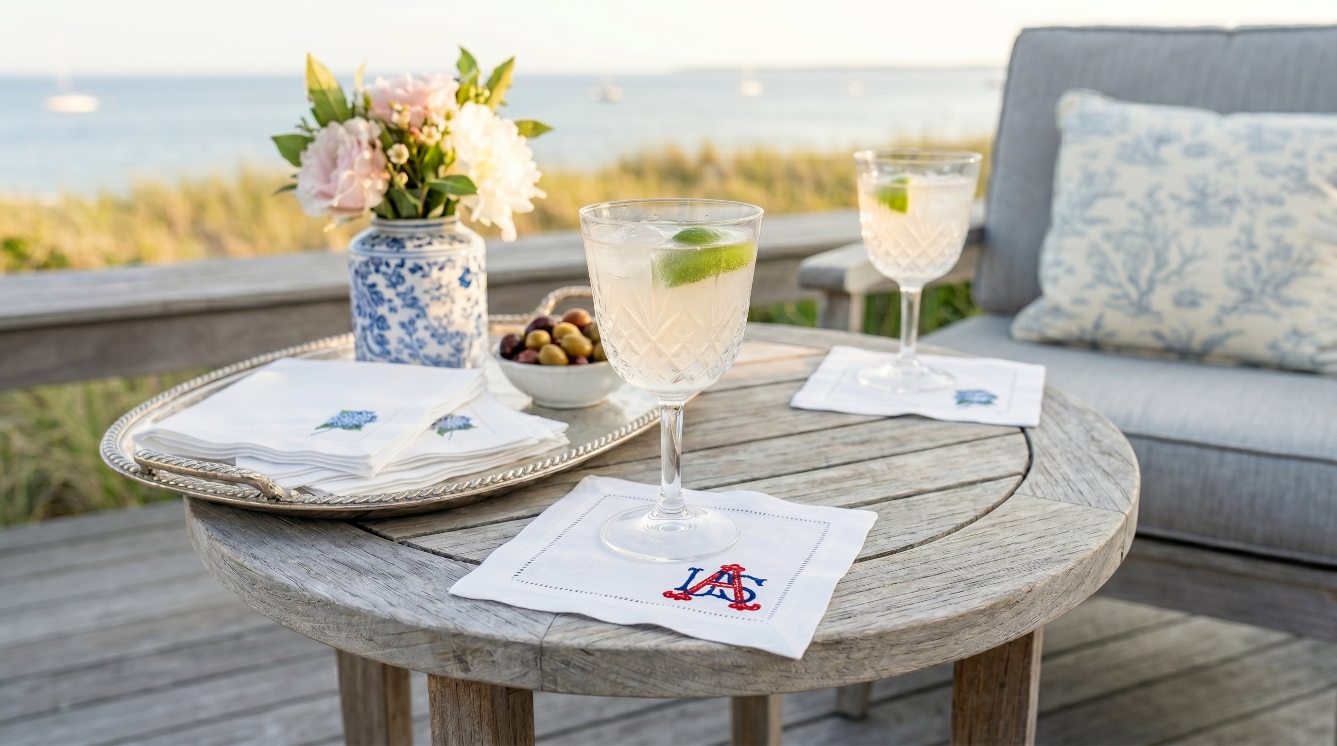 Outdoor setting with a wooden table, glasses of lemonade, and flowers on a deck. There are white linen embroidered cocktail napkins with hydrangeas and one with USA embroidered in the bottom right corner.
