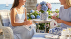 Two women sitting outdoors by a waterfront, enjoying a meal together with floral arrangements and a table setting. They are holding embroidered cocktail napkins with hydrangea and USA embroidered on them.