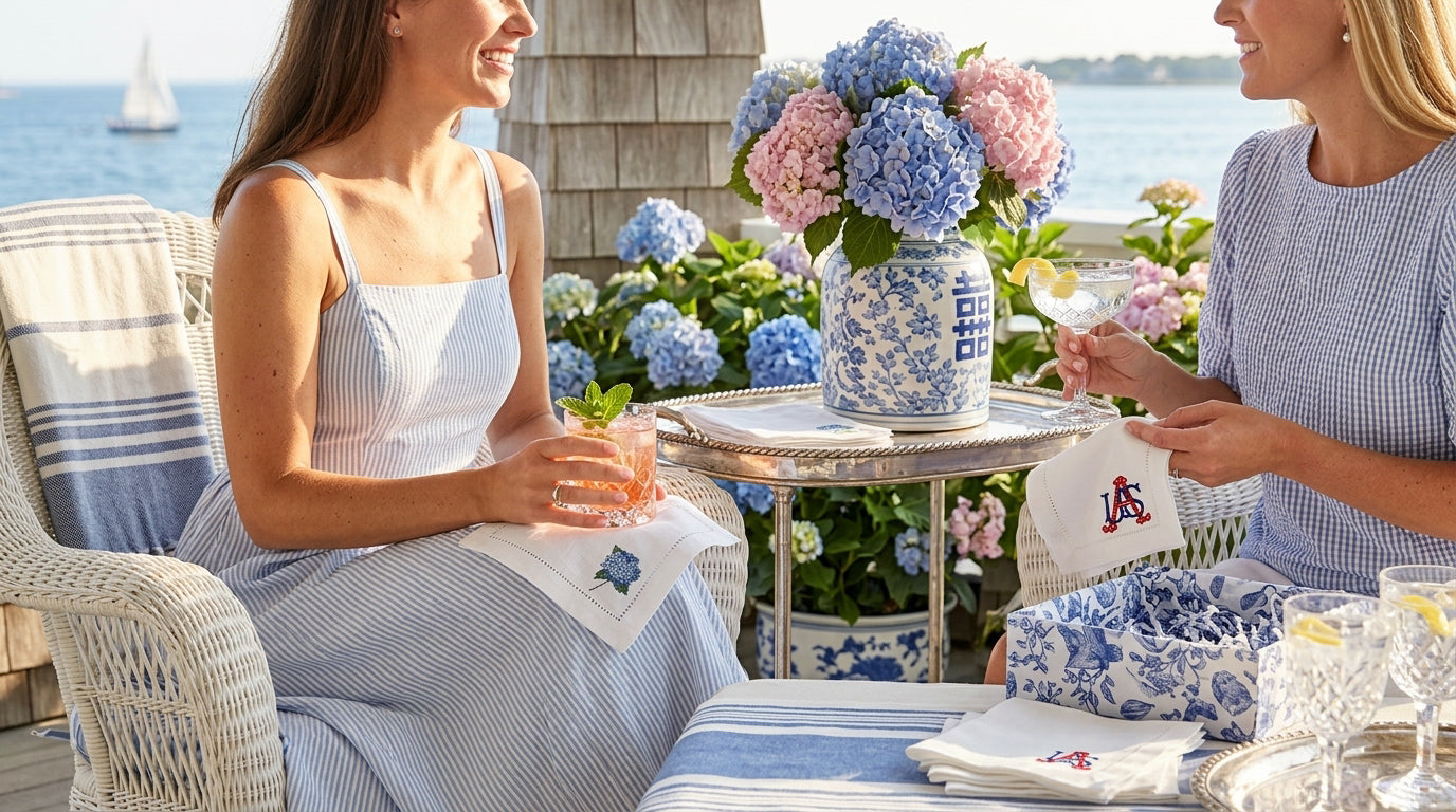 Two women sitting outdoors by a waterfront, enjoying a meal together with floral arrangements and a table setting. They are holding embroidered cocktail napkins with hydrangea and USA embroidered on them.