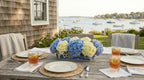 Outdoor dining table set with a floral centerpiece, glasses of iced tea, and views of a harbor.