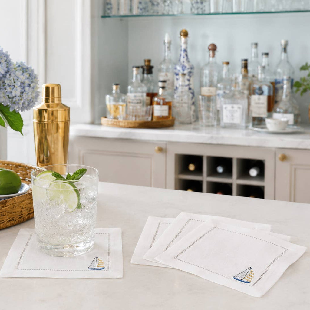 Four white linen hemstitched napkins set on a kitchen island with a cocktail on one. There is a sailboat in blue, white, and yellow stitched above the hemstitch in the right corner. There are hydrangeas and chinoiserie elements along with bar items in the room.
