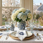 Elegant table setting with blue and white hydrangeas in a decorative vase. The table is set with monogrammed dinner and cocktail napkins.