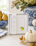 A boxed Sweet Humble Home set of 4 100% linen napkins with a golden pear embroidery design is displayed on a tabletop with blue-and-white ceramic decor, pears, a small pumpkin, and autumn leaves in a bright styled interior setting.