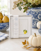 A boxed Sweet Humble Home set of 4 100% linen napkins with a golden pear embroidery design is displayed on a tabletop with blue-and-white ceramic decor, pears, a small pumpkin, and autumn leaves in a bright styled interior setting.