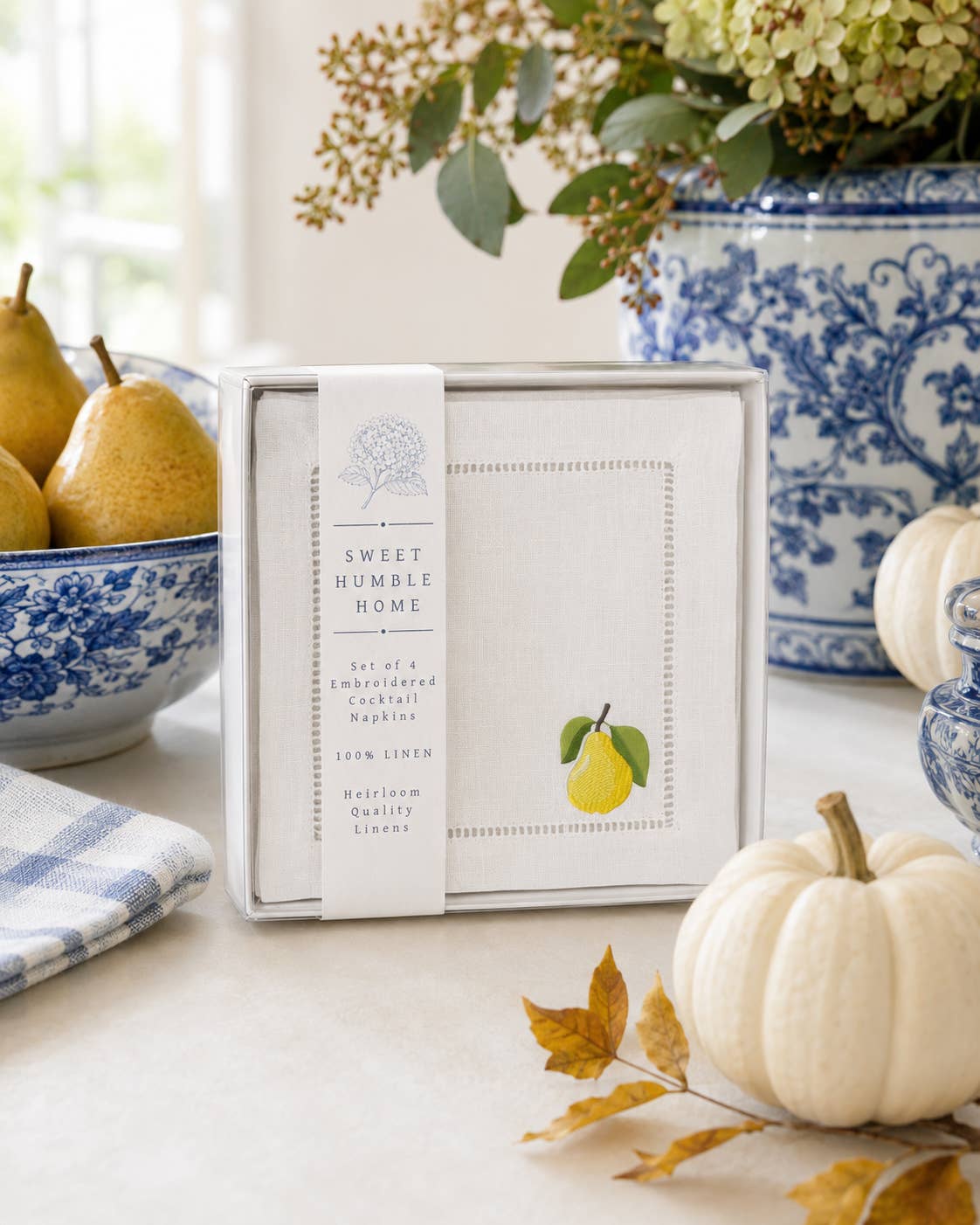 A boxed Sweet Humble Home set of 4 100% linen napkins with a golden pear embroidery design is displayed on a tabletop with blue-and-white ceramic decor, pears, a small pumpkin, and autumn leaves in a bright styled interior setting.