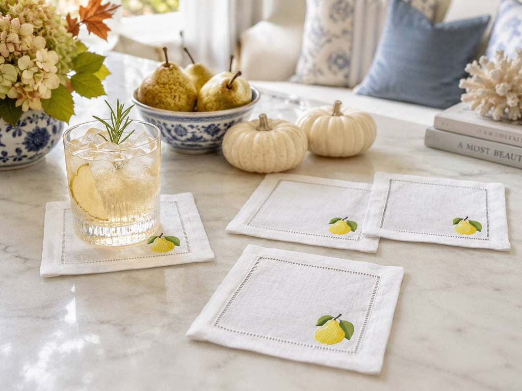 Four square white linen coasters with embroidered golden pear motifs are arranged on a marble table in a styled home setting, alongside a drink, pears, pumpkins, and decorative pillows.