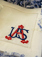 White linen cocktail napkin embroidered with a red and blue USA in a gift box with shredded chinoiserie style blue and white paper.