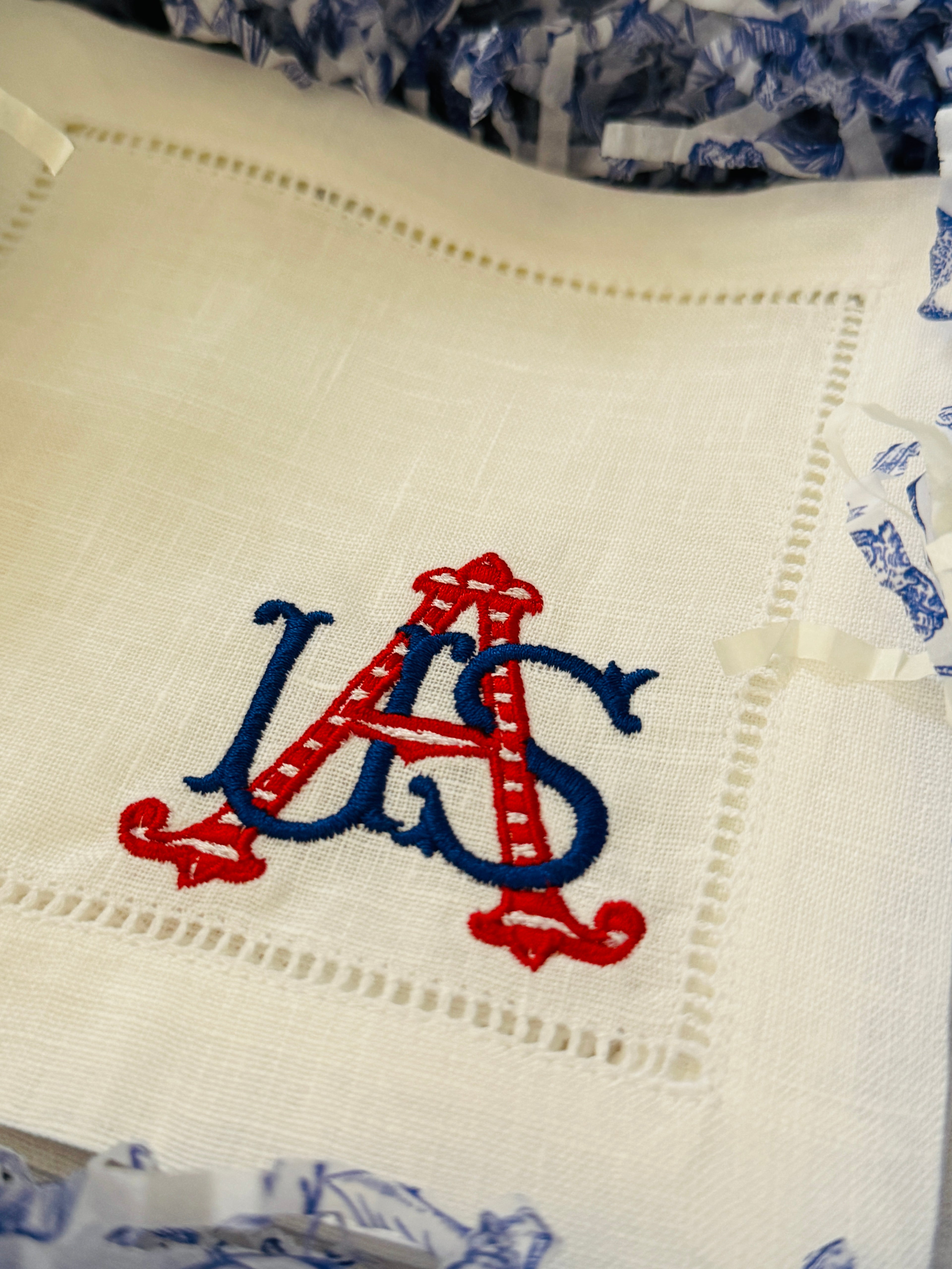 White linen cocktail napkin embroidered with a red and blue USA in a gift box with shredded chinoiserie style blue and white paper.