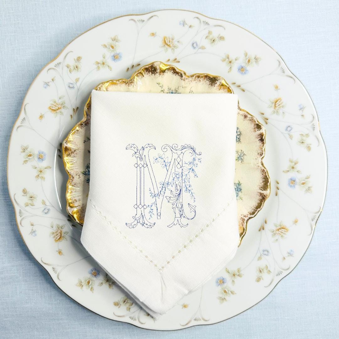 Vintage Floral M stitched in blue on a white napkin on top of mismatched china dinnerware.