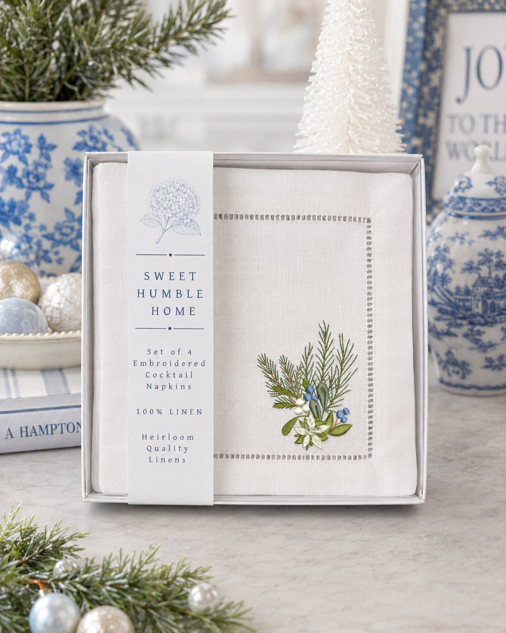 A boxed set of embroidered cocktail napkins from Sweet Humble Home, displayed in a gift box with a label. The napkins are white linen with blue and green botanical embroidery, and the scene is styled with blue-and-white ceramic vases, pine branches, and a white Christmas tree in a bright indoor setting.