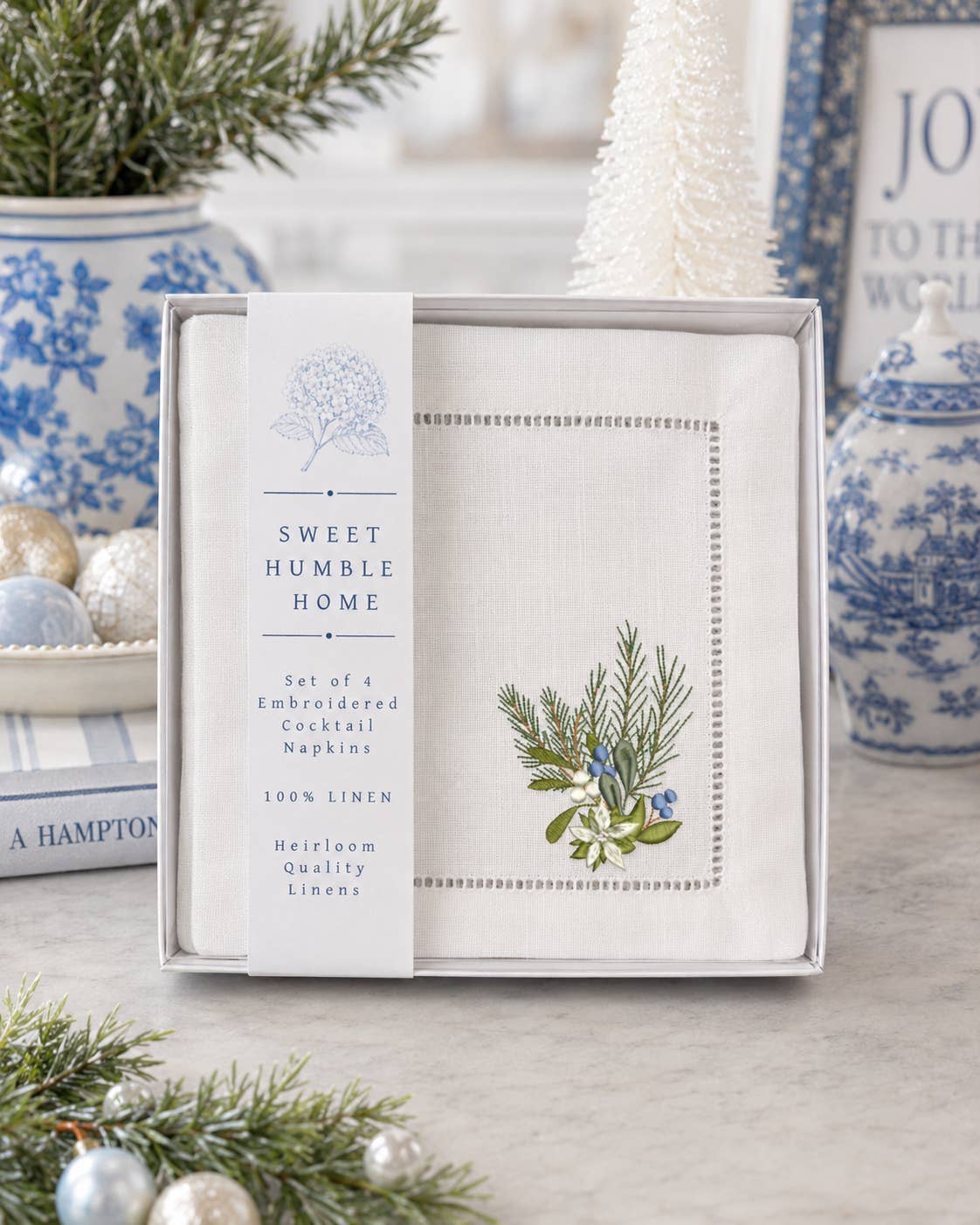 A boxed set of embroidered cocktail napkins from Sweet Humble Home, displayed in a gift box with a label. The napkins are white linen with blue and green botanical embroidery, and the scene is styled with blue-and-white ceramic vases, pine branches, and a white Christmas tree in a bright indoor setting.