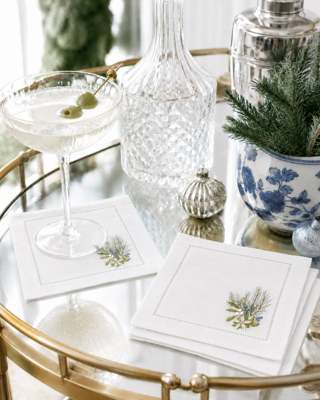 White embroidered napkins with a botanical design, styled on a gold bar cart next to a cocktail glass, a glass decanter, and holiday decorations.