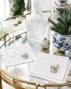 White embroidered napkins with a botanical design, styled on a gold bar cart next to a cocktail glass, a glass decanter, and holiday decorations.
