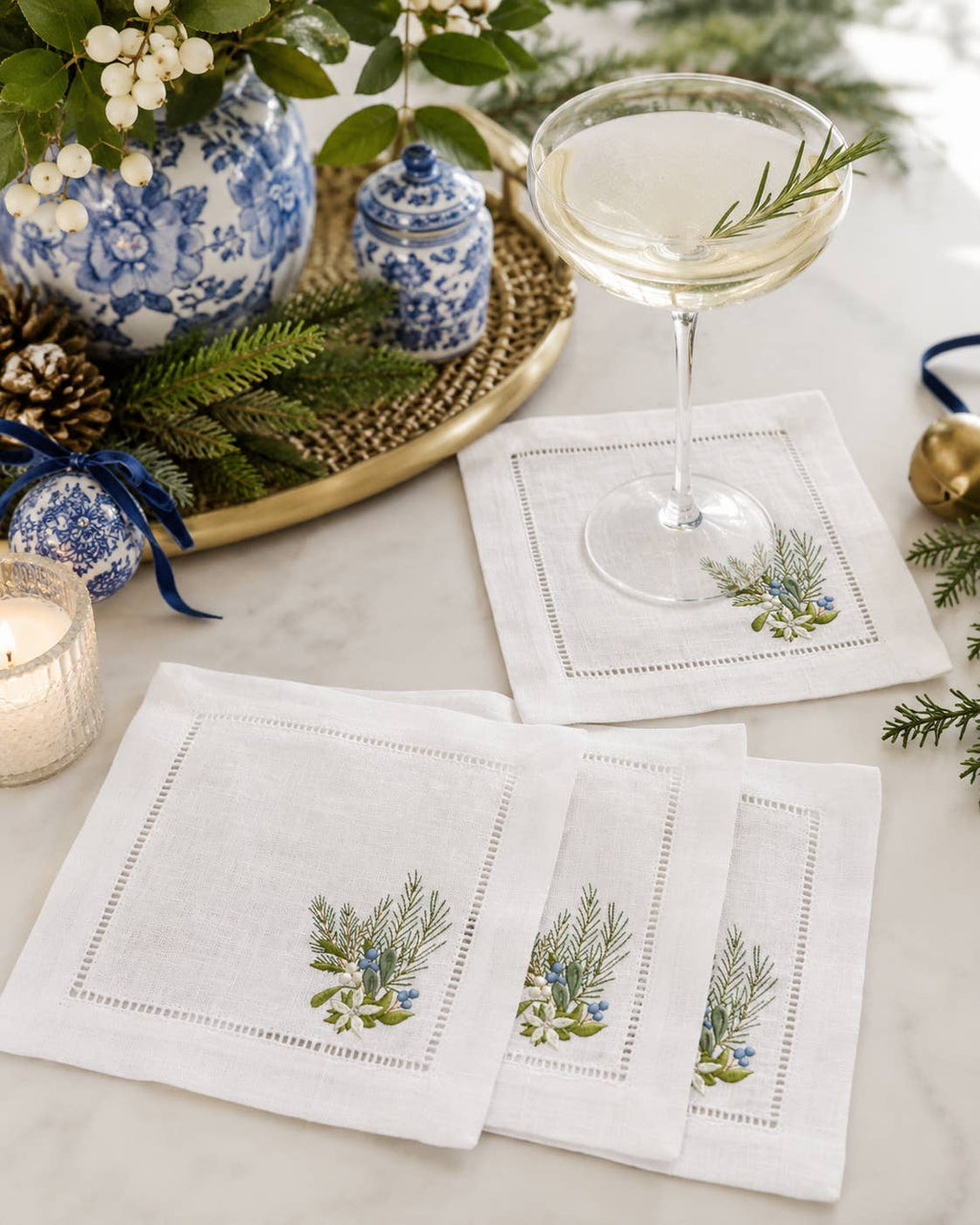 A set of white linen napkins with embroidered green and blue floral motifs, styled on a marble tabletop alongside a cocktail glass, blue-and-white ceramic vases, pine branches, and a candle.