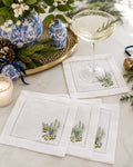 A set of white linen napkins with embroidered green and blue floral motifs, styled on a marble tabletop alongside a cocktail glass, blue-and-white ceramic vases, pine branches, and a candle.