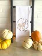A white tea towel with the outline of a brown turkey embroidered with the words inside of the turkey: Pour Some Gravy on Me. The towel is set on a countertop black towel ladder with mini pumpkins to the left and right.
