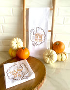 Two white tea towels each embroidered with the outline of a turkey in brown with the words: Leftovers are for Quitters embroidered inside the turkey on the right which is hanging on a towel ladder on a white marble counter,  and Pour Some Gravy on me on the towel on the left that is sitting folded on round wooden tray. There are colorful little pumpkins to the left and right of the towels as well.