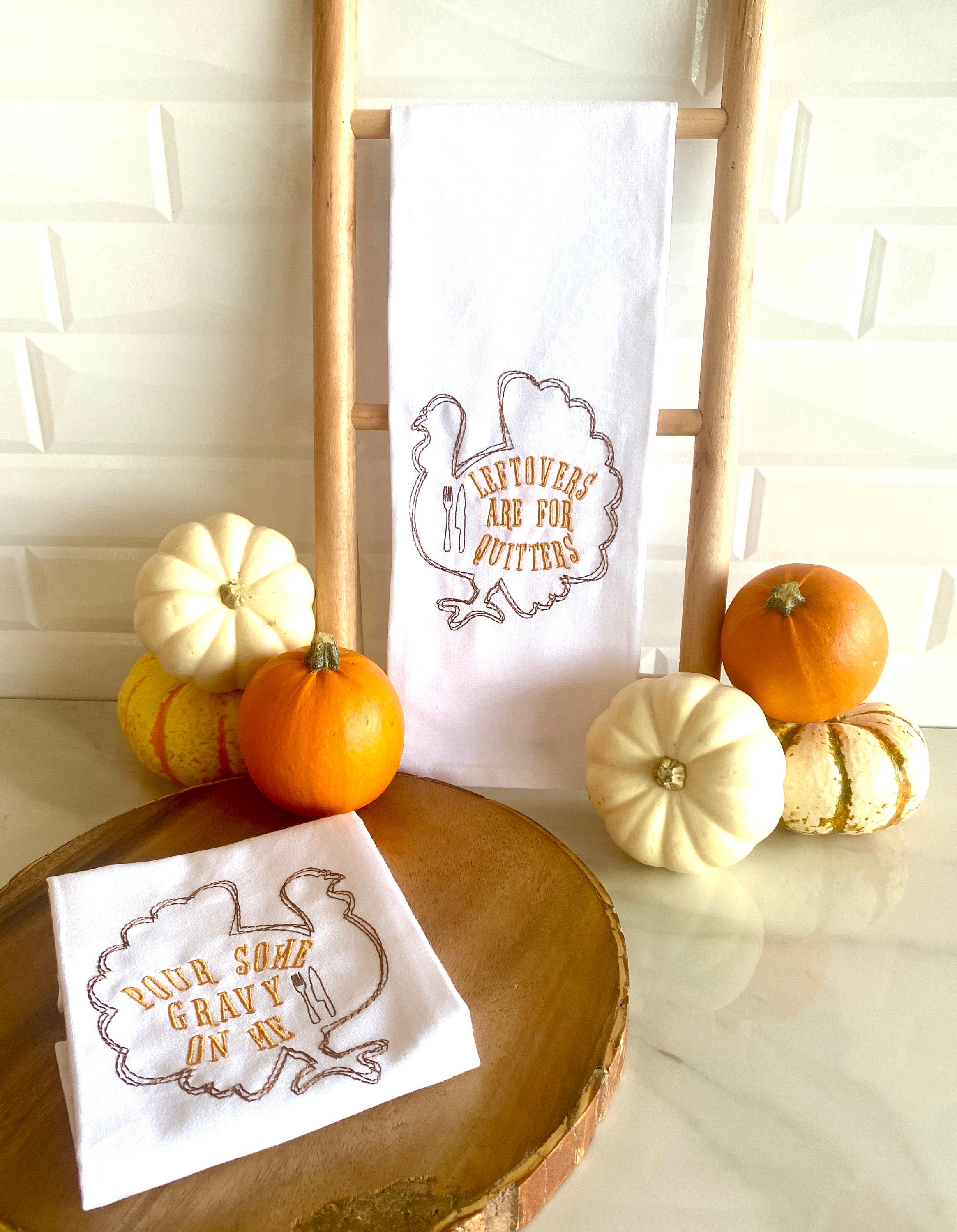 Two white tea towels each embroidered with the outline of a turkey in brown with the words: Leftovers are for Quitters embroidered inside the turkey on the right which is hanging on a towel ladder on a white marble counter,  and Pour Some Gravy on me on the towel on the left that is sitting folded on round wooden tray. There are colorful little pumpkins to the left and right of the towels as well.