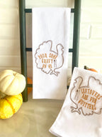 Two white tea towels each embroidered with the outline of a turkey in brown with the words: Leftovers are for Quitters embroidered inside the turkey on the right and Pour Some Gravy on me on the towel on the left. The towels are set on a white marble countertop with colorful little pumpkins set along the top.