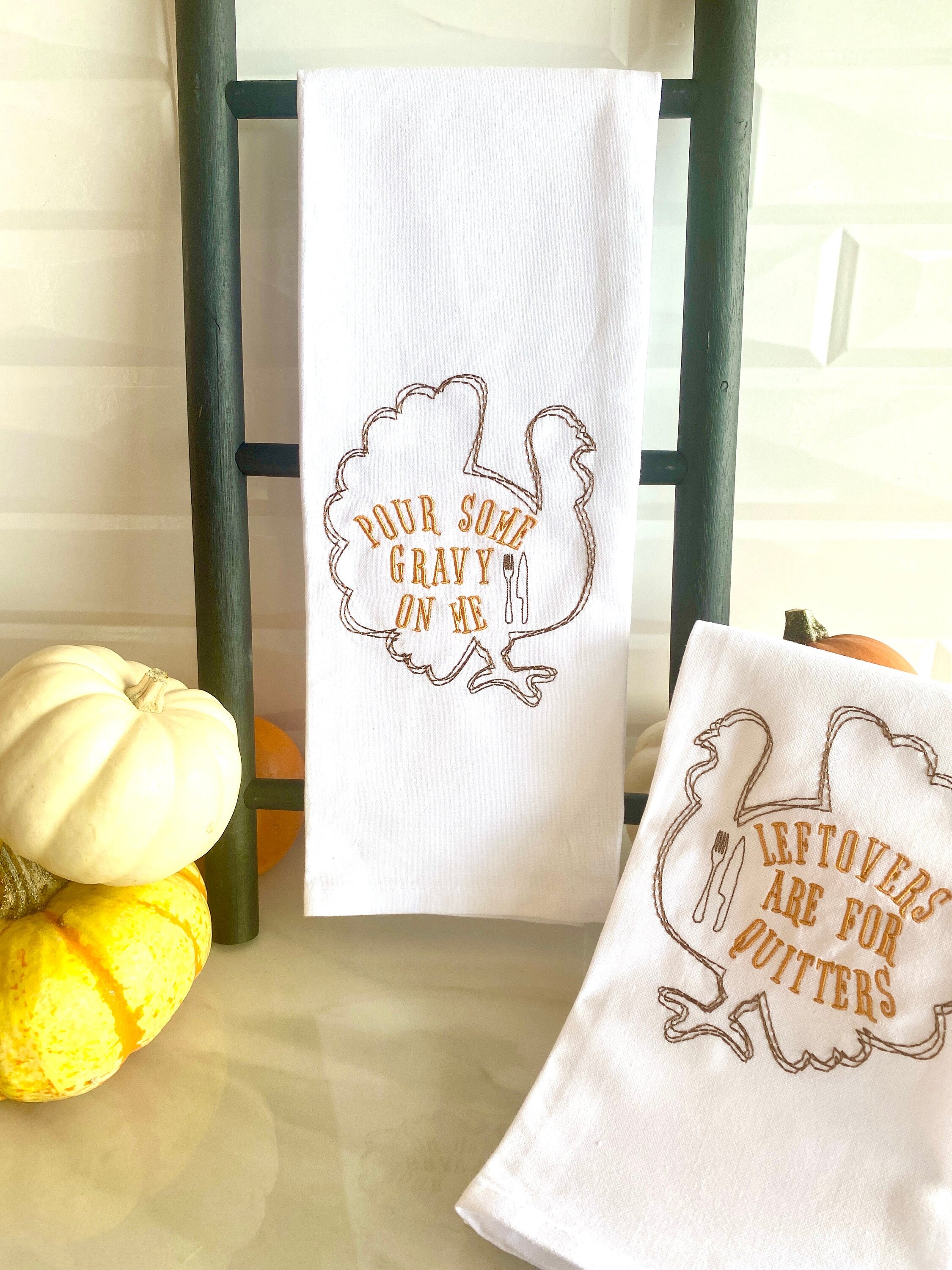 Two white tea towels each embroidered with the outline of a turkey in brown with the words: Leftovers are for Quitters embroidered inside the turkey on the right and Pour Some Gravy on me on the towel on the left. The towels are set on a white marble countertop with colorful little pumpkins set along the top.