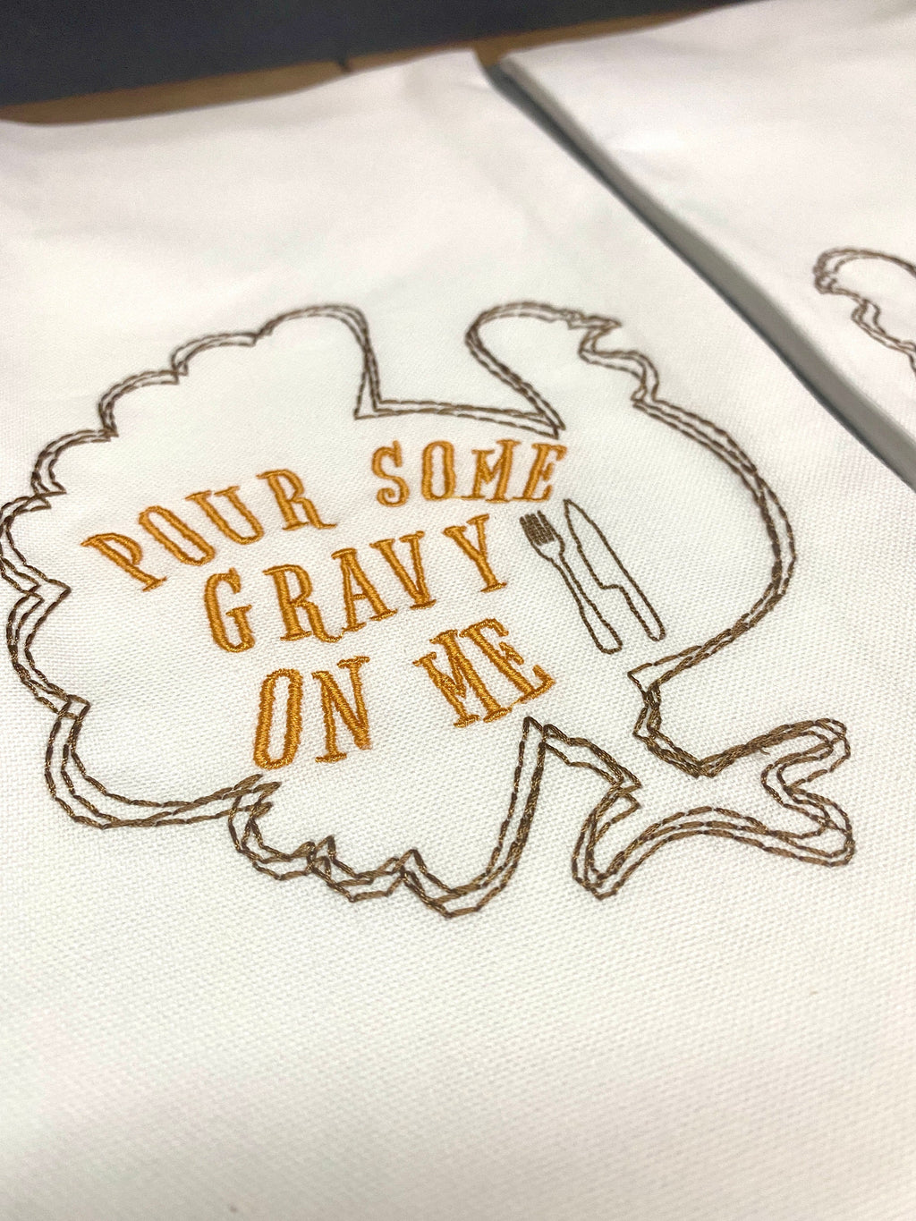 A white tea towel with the outline of a brown turkey embroidered with the words inside of the turkey: Pour Some Gravy on Me.