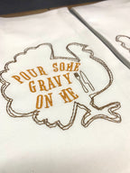 A white tea towel with the outline of a brown turkey embroidered with the words inside of the turkey: Pour Some Gravy on Me.