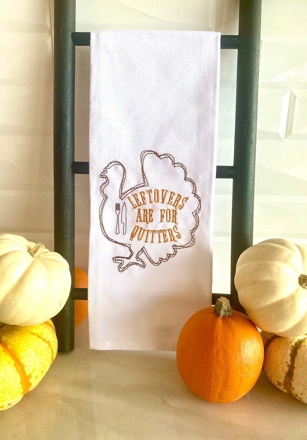 A white tea towel embroidered with the outline of a turkey in brown with the words: Leftovers are for Quitters embroidered inside the turkey. The towel is hanging on a black towel countertop ladder on a white marble counter and white subway tile backsplash with colorful little pumpkins to the right and the left.