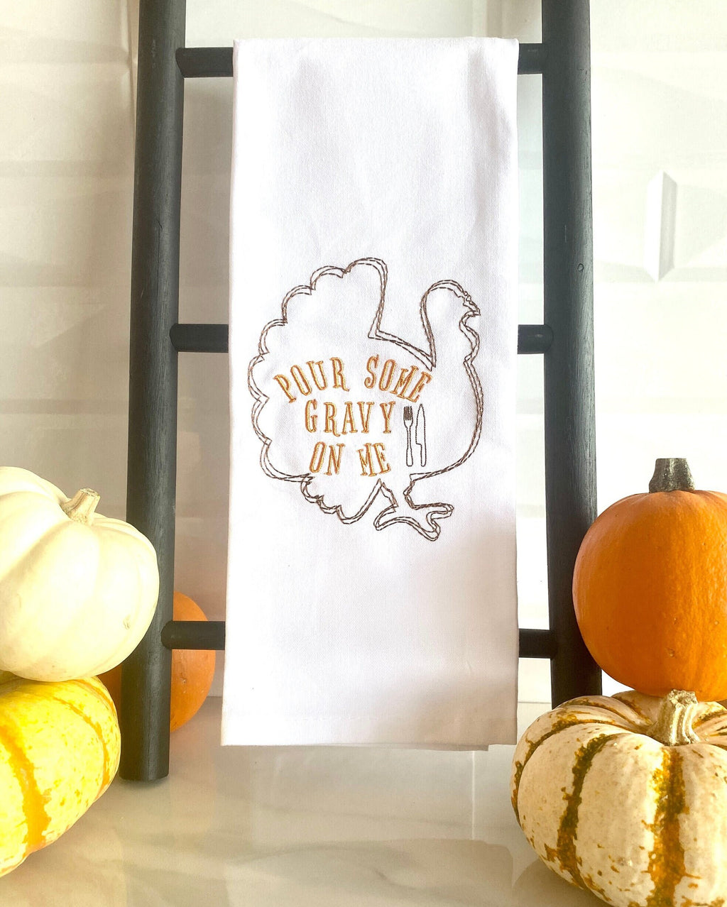A white tea towel with the outline of a brown turkey embroidered with the words inside of the turkey: Pour Some Gravy on Me. The towel is set on a countertop black towel ladder with mini pumpkins to the left and right.
