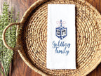 A dreidel in different shades of blue with a pink bow embroidered on a white cotton tea towel set on a basket with custom embroidery chosen as an option with the name: Goldberg Family. It is set in a basket on a table with greens to the left side.