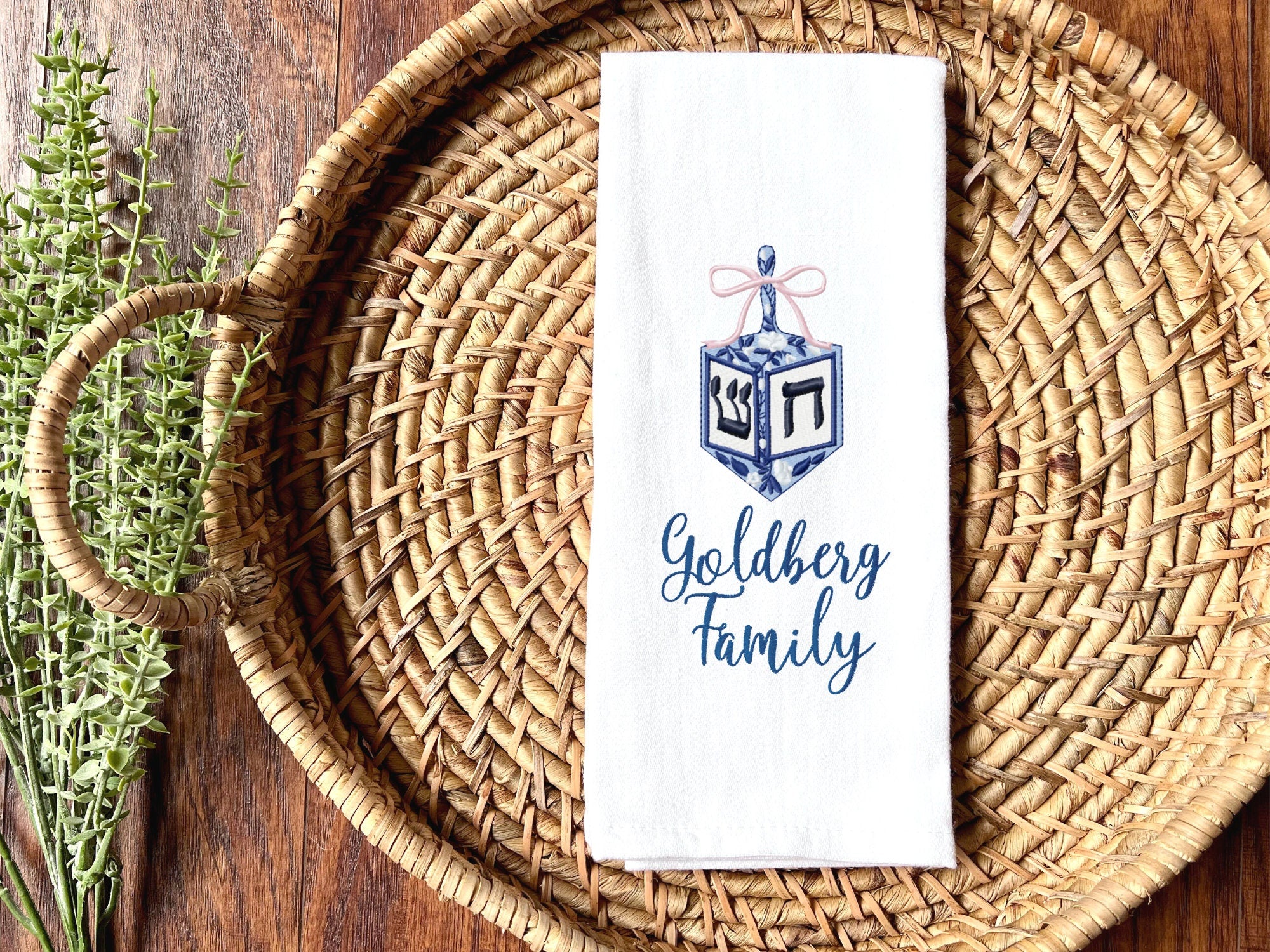 A dreidel in different shades of blue with a pink bow embroidered on a white cotton tea towel set on a basket with custom embroidery chosen as an option with the name: Goldberg Family. It is set in a basket on a table with greens to the left side.