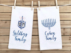 A photo of two white tea towel one with a dreidel in different shades of blue with a pink bow embroidered on a white cotton tea towel and one with a menorah in different shades of blue on a white cotton tea towel. Both are personalized with custom family names. They are set on a wood slatted background.